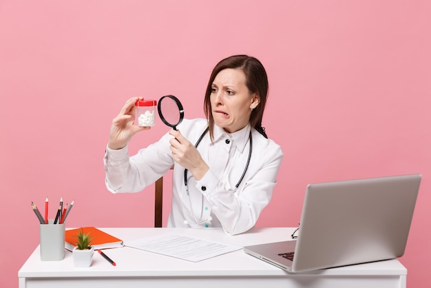 Femme médecin s'asseoir au bureau travailler sur ordinateur avec document médical tenir des pilules à l'hôpital isolé sur fond de mur rose pastel. femme en robe médicale lunettes stéthoscope. concept de médecine de santé.