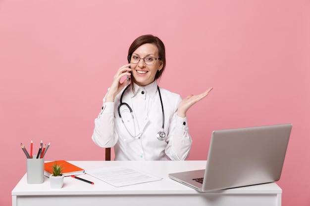 Femme médecin s'asseoir au bureau travailler sur ordinateur avec document médical tenir téléphone portable à l'hôpital isolé sur fond rose pastel. femme en robe médicale lunettes stéthoscope. concept de médecine de santé.