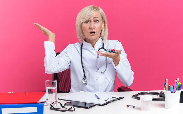 Femme médecin slave adulte anxieuse en robe médicale avec stéthoscope assis au bureau avec des outils de bureau tenant des comprimés de médicaments sous blister isolé sur fond rose avec espace de copie