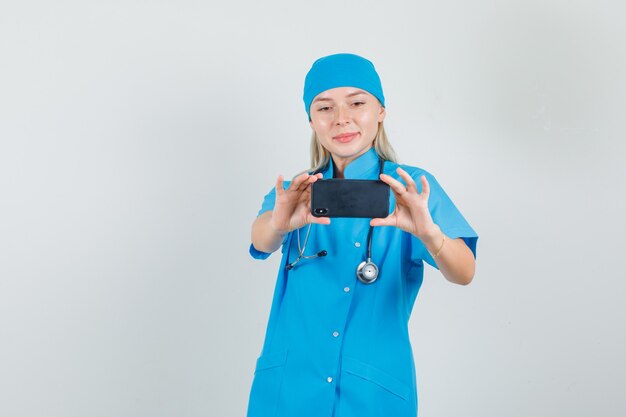 Femme médecin souriant et prenant des photos sur smartphone en uniforme bleu