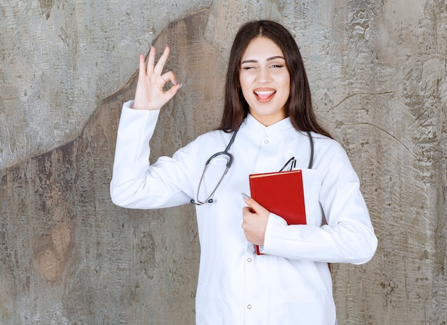 Femme médecin souriante geste OK et tenant un livre sur un mur gris