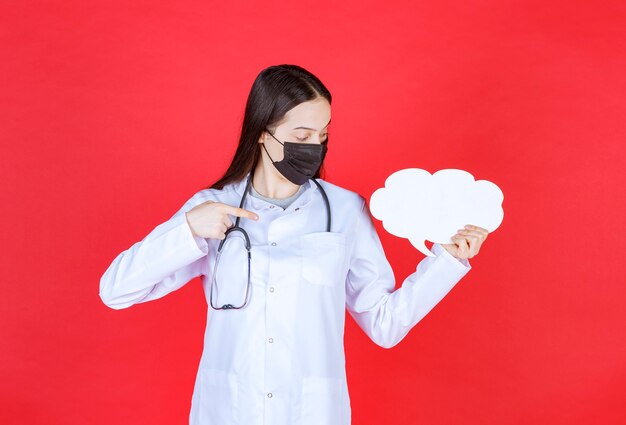 Femme médecin avec stéthoscope et masque noir tenant un bureau d'informations vide en forme de nuage.