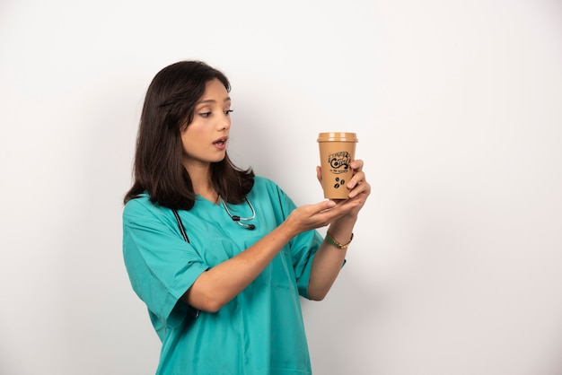 Femme médecin avec stéthoscope à la recherche de café sur fond blanc.