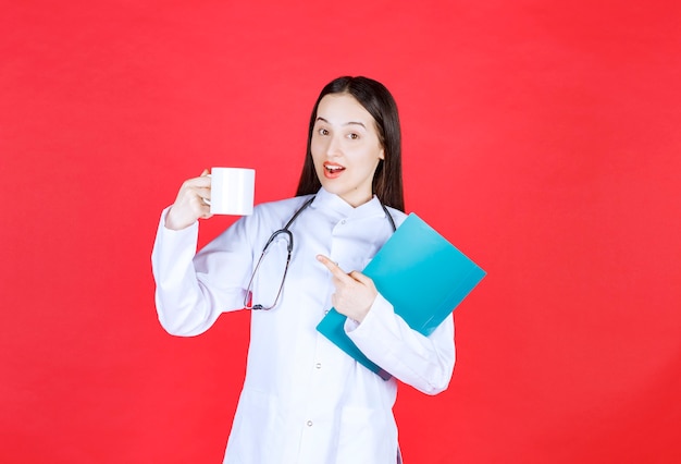 Femme médecin avec stéthoscope tenant une tasse de boisson et un dossier d'historique du patient.