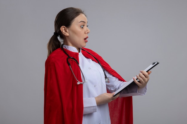 Femme médecin avec stéthoscope en uniforme médical blanc et cape de super-héros rouge montrant le presse-papiers avec des pages vierges à la surprise et surpris debout sur un mur blanc