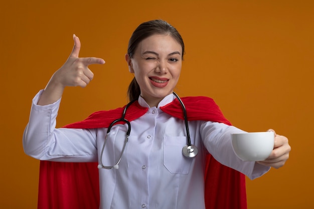 Femme médecin avec stéthoscope en uniforme médical blanc et cape de super-héros rouge montrant une tasse de café pointant avec le doigt dessus en souriant et en clignant de l'œil debout sur un mur orange