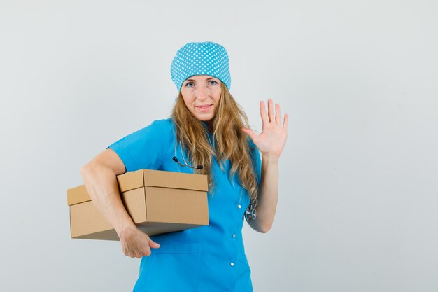 Femme médecin tenant une boîte en carton, agitant la main en uniforme bleu et à la recherche de joie.