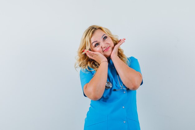 Femme médecin tenant les mains sur le menton en uniforme bleu et à la recherche de plaisir.