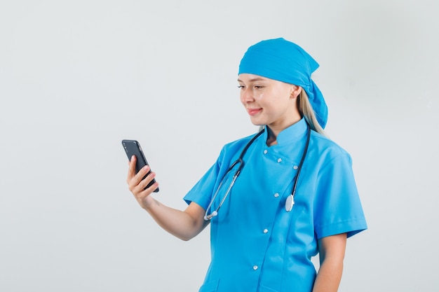 Femme médecin tenant et regardant smartphone en uniforme bleu et à la recherche de bonne humeur.