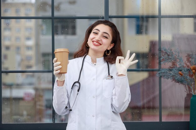 Femme médecin tenant une tasse de café et geste OK Photo de haute qualité