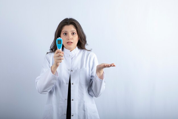 Femme médecin avec thermomètre debout sur un mur blanc.