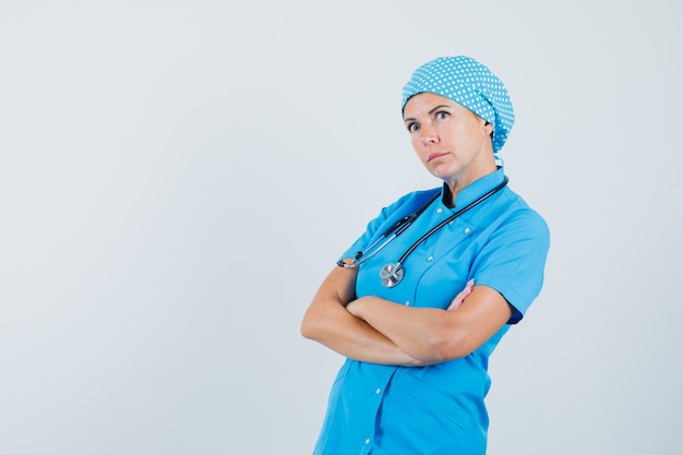 Femme médecin en uniforme bleu debout avec les bras croisés et regardant pensif, vue de face.