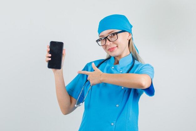 Femme médecin en uniforme bleu, lunettes pointant le doigt sur smartphone et à la bonne humeur