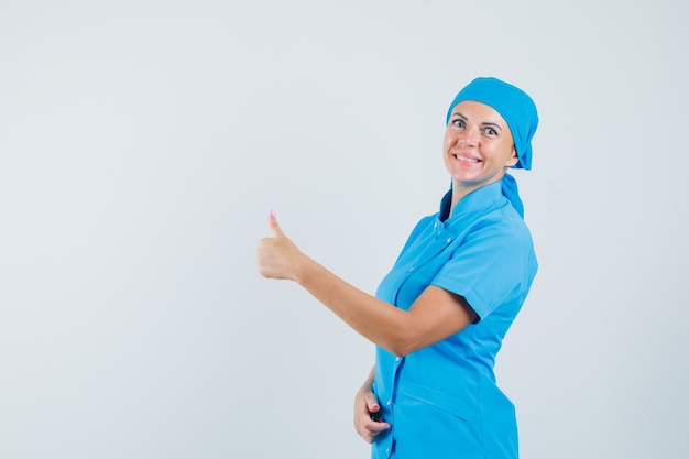 Femme médecin en uniforme bleu montrant le pouce vers le haut et à la joyeuse vue de face.