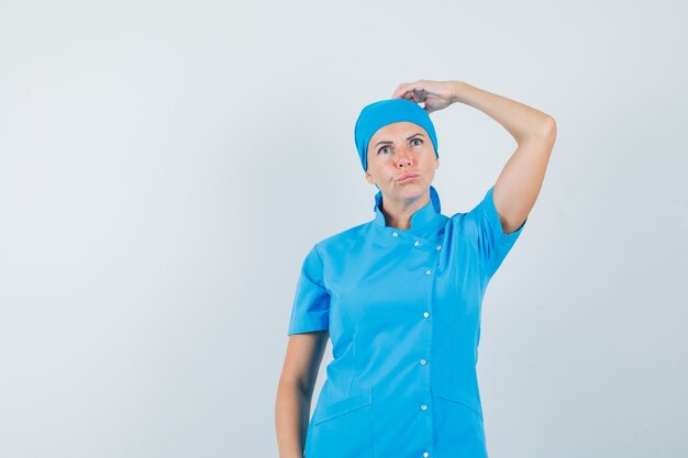 Femme médecin en uniforme bleu se gratter la tête et regardant pensif, vue de face.