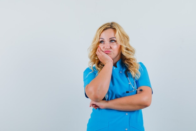 Femme médecin en uniforme bleu se penchant sur sa main tout en regardant ailleurs et à l'insatisfaction