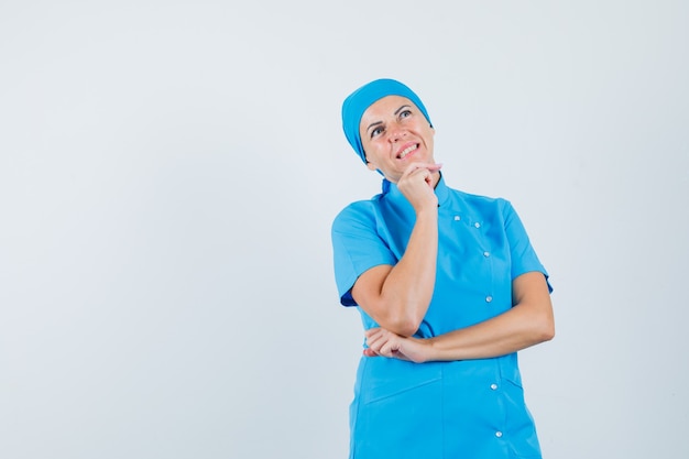 Femme médecin en uniforme bleu soutenant le menton sur place et à la vue de face, hésitante.