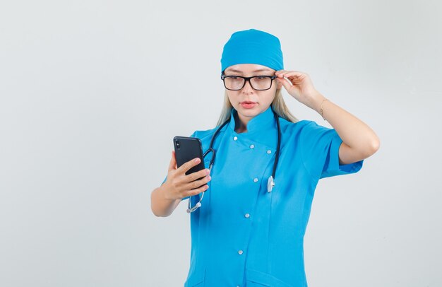 Femme médecin en uniforme bleu tenant le smartphone avec la main sur des lunettes et à la recherche de occupé