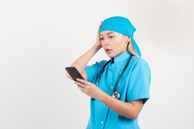 Femme médecin en uniforme bleu tenant le smartphone avec la main sur la tête et à la confusion