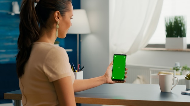 Femme de médias sociaux tenant un téléphone à écran tactile avec une clé de chrominance d'écran vert simulée, assise sur un bureau moderne. femme de race blanche travaillant sur une application en ligne à l'aide d'un smartphone avec écran isolé