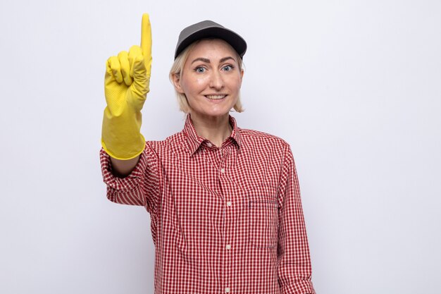 Femme de ménage en chemise à carreaux et casquette portant des gants en caoutchouc à sourire montrant l'index