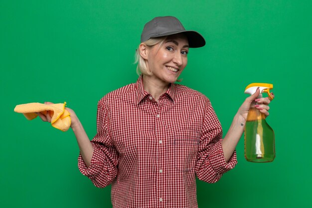 Femme de ménage en chemise à carreaux et casquette tenant un chiffon et un spray de nettoyage à la recherche de sourire confiant prêt pour le nettoyage