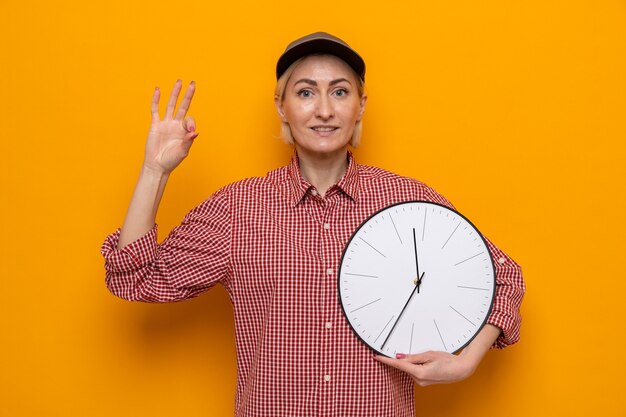 Femme de ménage en chemise à carreaux et casquette tenant une horloge à la recherche d'un sourire confiant montrant un signe ok