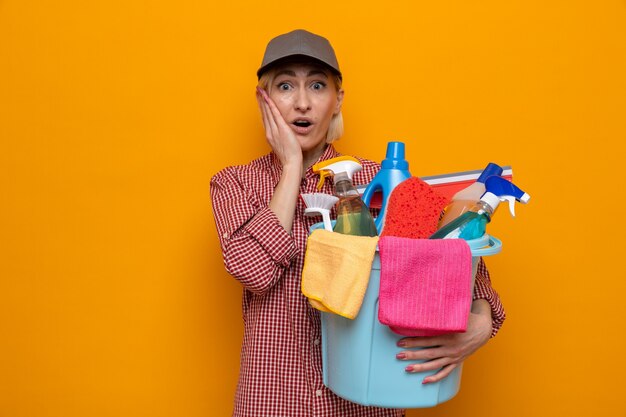 Femme de ménage en chemise à carreaux et casquette tenant un seau avec des outils de nettoyage regardant la caméra étonnée et surprise debout sur fond orange