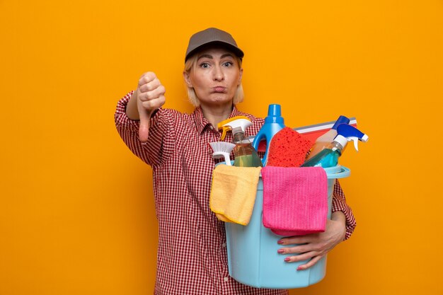 Femme de ménage insatisfaite en chemise à carreaux et casquette tenant un seau avec des outils de nettoyage regardant les pouces vers le bas