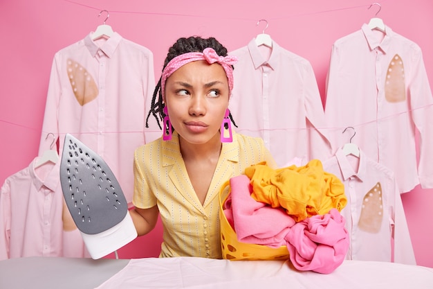 La femme de ménage pensive à la peau sombre et mécontente regarde pensivement loin tient une pile sur du linge déplié et du fer électrique à vapeur va caresser le linge porte un bandeau tenue décontractée travaille dans le lavoir