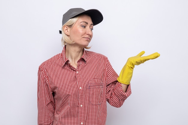 Femme de ménage sérieuse en chemise à carreaux et casquette portant des gants en caoutchouc regardant son bras présentant quelque chose