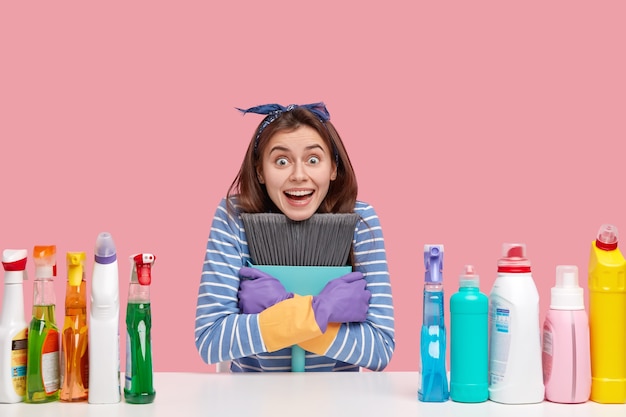 Photo gratuite une femme de ménage souriante a une expression étonnée, un large sourire, porte le balai de près, croise les mains sur la poitrine, porte des gants en caoutchouc, heureux d'avoir une journée de congé