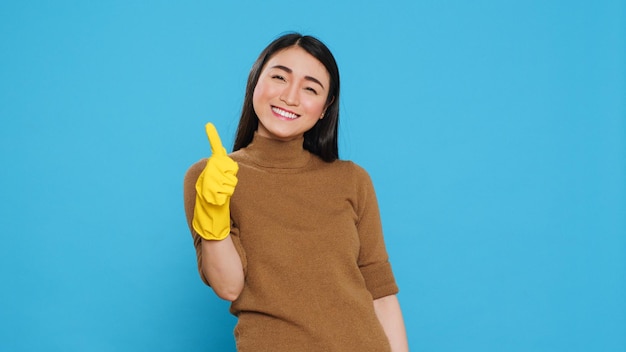 Femme de ménage souriante portant des gants en caoutchouc jaune tout en faisant un geste correct après avoir fini de nettoyer la maison du client, posant en studio. La femme de ménage gaie est responsable de fournir des services de nettoyage
