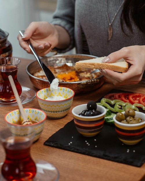 Une femme met de la crème sur une tranche de pain autour d’une table de petit-déjeuner contenant beaucoup d’aliments.