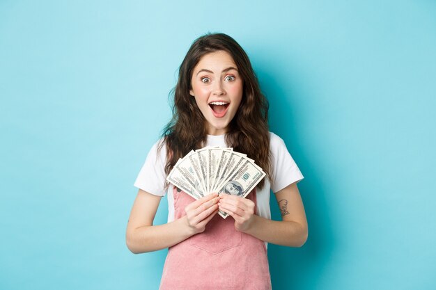 Femme mignonne excitée gagnant de l'argent, tenant des billets d'un dollar et souriante étonnée, a obtenu un crédit rapide, debout sur fond bleu.