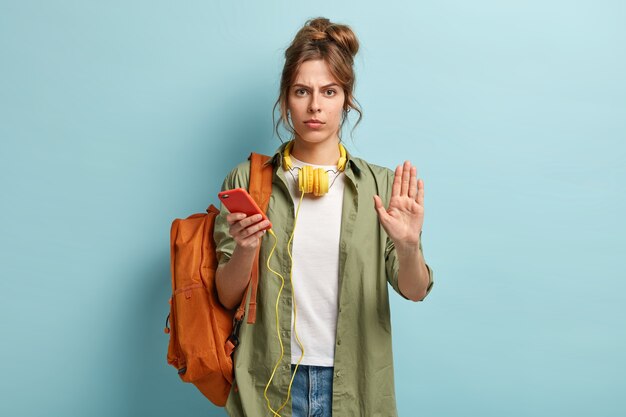 Une femme mignonne sérieuse passe du temps à l'intérieur avec un gadget moderne, montre un geste d'arrêt, demande à ralentir, voyage avec de la musique et un sac à dos