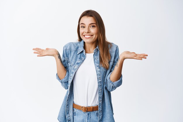 Femme mignonne souriante haussant les épaules et souriante, guity comme accusé, debout contre un mur blanc dans des vêtements décontractés