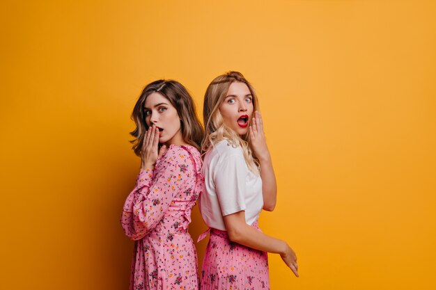 Femme mignonne surprise en t-shirt blanc debout à côté d'un ami. Photo intérieure de joyeuses filles caucasiennes exprimant leur étonnement.