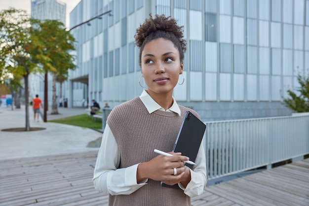 Une femme millénaire réfléchie aux cheveux bouclés noirs se promène dans la rue de la ville moderne tient un stylet tactile portable profite de la vue sur les bâtiments urbains utilise les technologies Femme avec tablette numérique