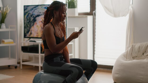 Femme mince assise sur un ballon de fitness écrivant un message au téléphone pendant l'entraînement du matin dans le salon