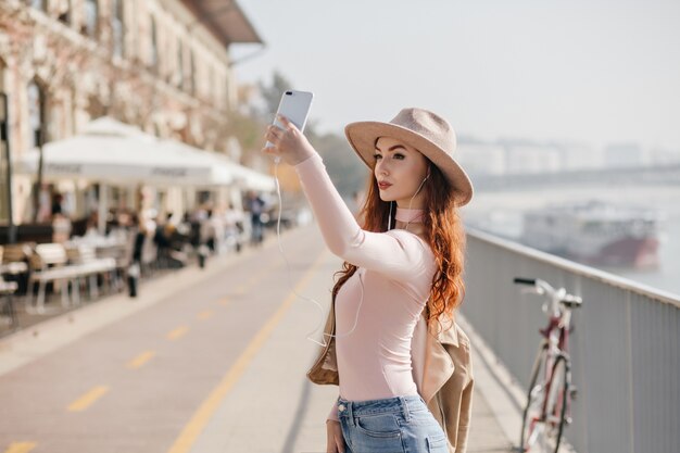 Femme mince au chapeau beige à la mode faisant selfie avec une expression de visage grave