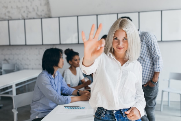 Femme mince aux cheveux courts blonds montrant un signe correct après une négociation réussie. Portrait d'employé de bureau asiatique posant avec un collègue africain lors de la conférence avec une femme blonde.