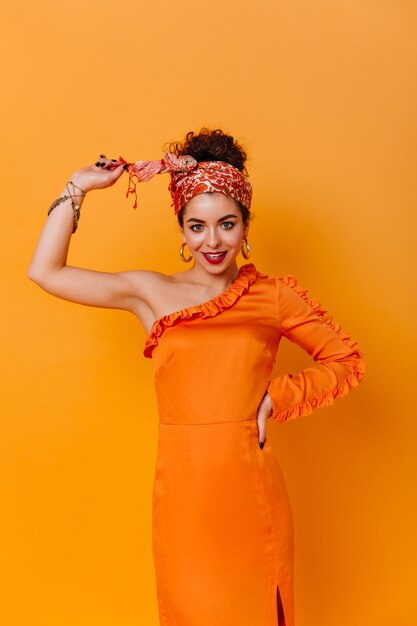 Une femme mince dans une élégante robe orange et un bandeau de style africain sourit doucement contre l'espace orange.