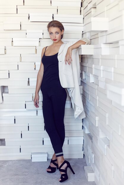 Femme mince dans un vêtement noir posant près d'un mur blanc.