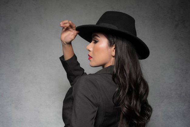 Une femme mince sexy portant un costume noir à la mode sans soutien-gorge et chapeau noir est une mode sexy aux tons sombres qui pose en studio isolé sur fond gris.