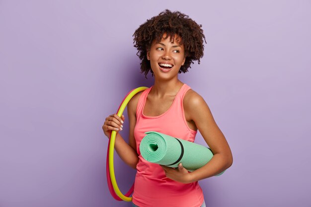 Femme mince sportive avec une peau foncée saine, coiffure afro, exercices avec cerceau, porte tapis roulé, vêtue d'un gilet rose, a le sourire à pleines dents