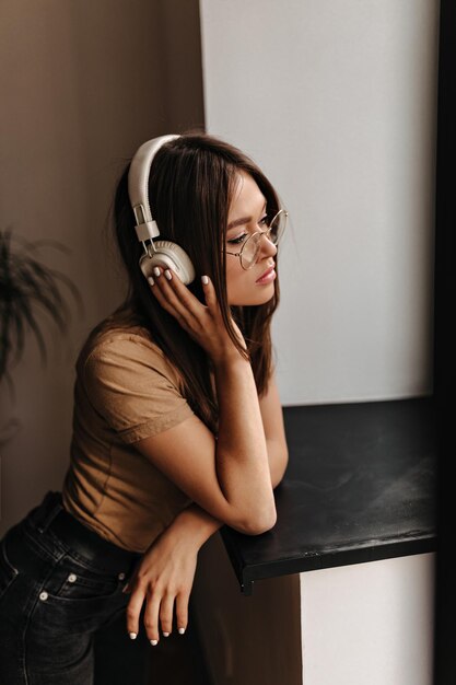 Femme mince en t-shirt beige et jeans noirs écoute de la musique avec des écouteurs appuyés sur le rebord de la fenêtre