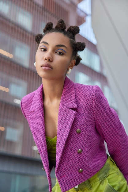 Photo gratuite une femme à la mode attentive avec une coiffure en chignon regarde sérieusement la caméra vêtue d'une veste rose élégante pose sur fond de ville a un maquillage vif et lumineux. style de personnes et concept de mode