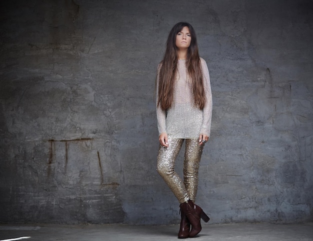 Femme à la mode aux longs cheveux bruns en costume brillant posant sur un mur gris.