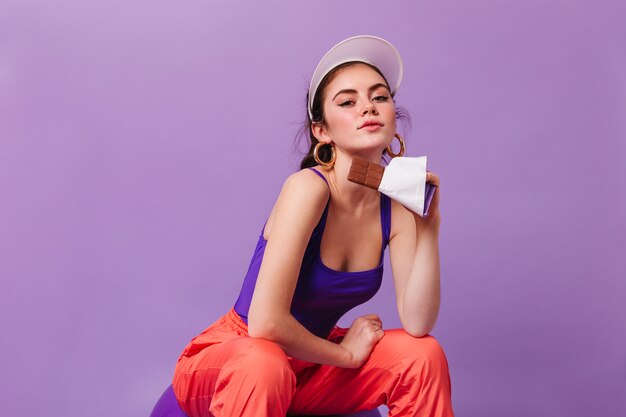 Femme à la mode avec des boucles d'oreilles massives et en cap à l'avant et tenant la barre de chocolat sur le mur violet